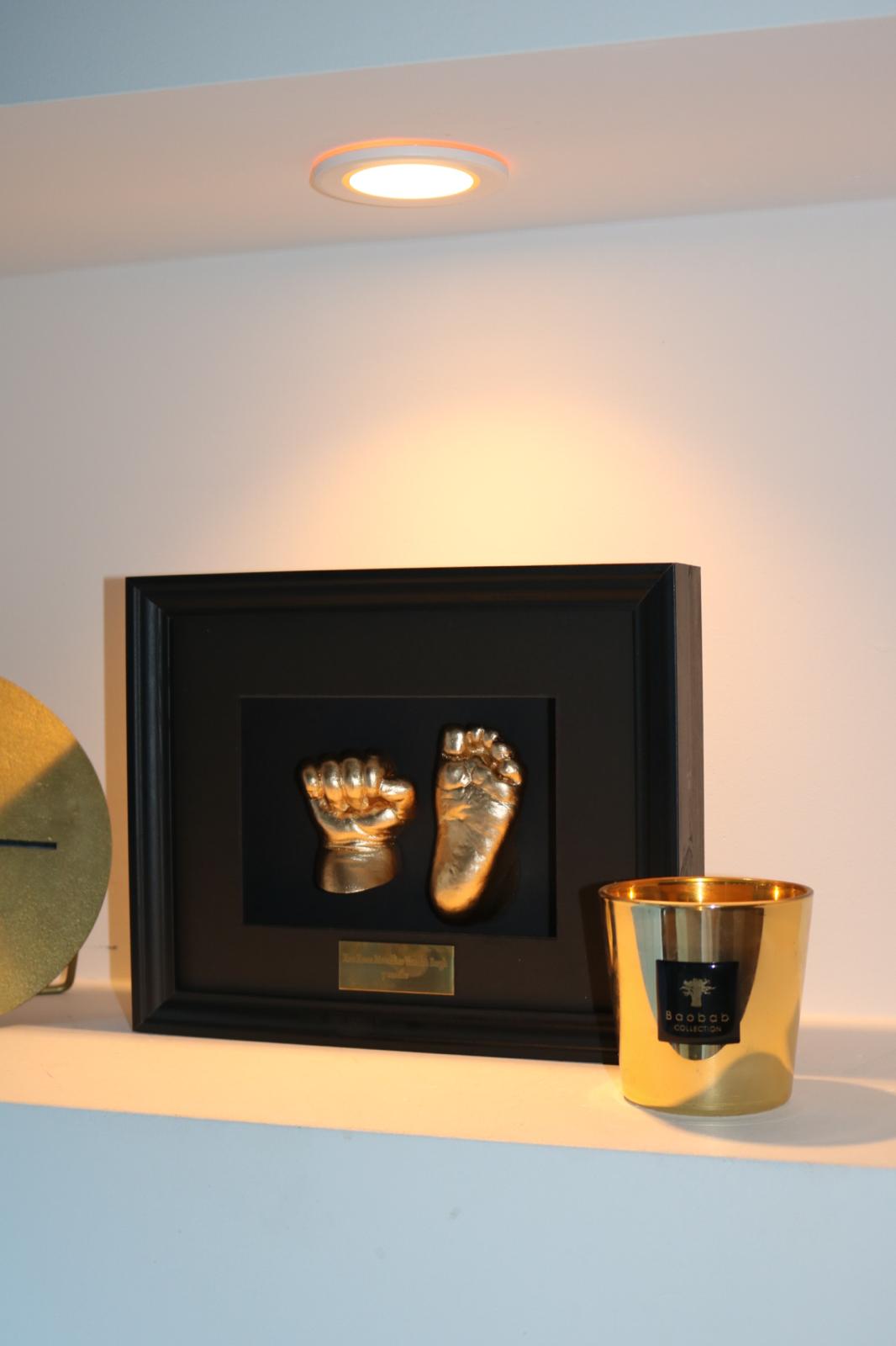 Gouden afdrukken van een babyhandje en voetje in een zwarte lijst, op een verlichte plank naast een gouden kaars