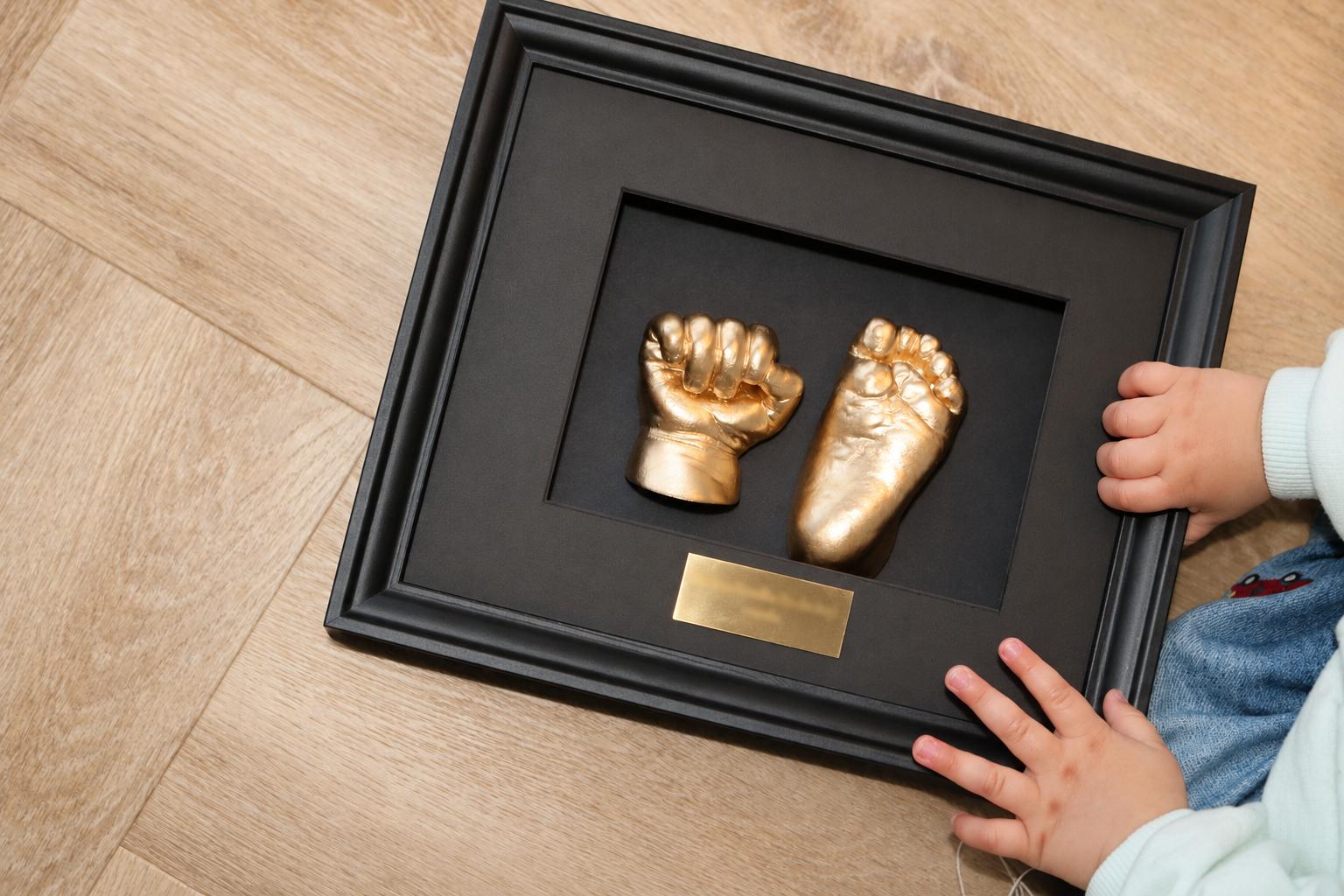 Een kindje bekijkt de gouden afdrukken van een handje en voetje in een zwarte 3D-lijst
